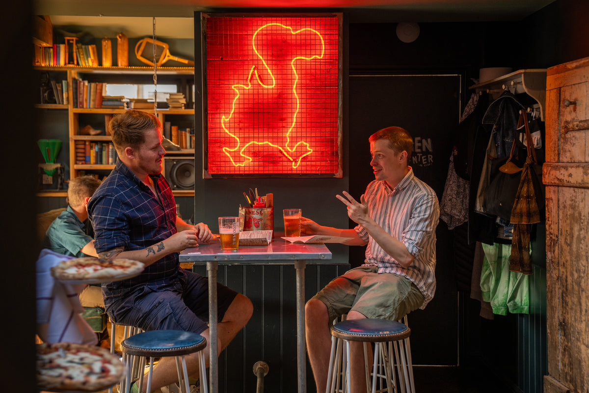 Two friends chatting over drinks at a high table beneath a red neon dodo sign in a Dodo pub