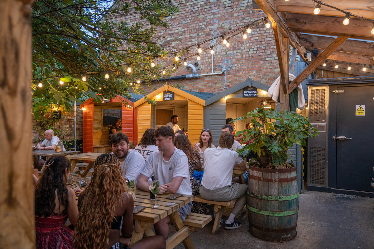 Busy beer garden with festoon lights, wooden tables and colourful beach-hut booths