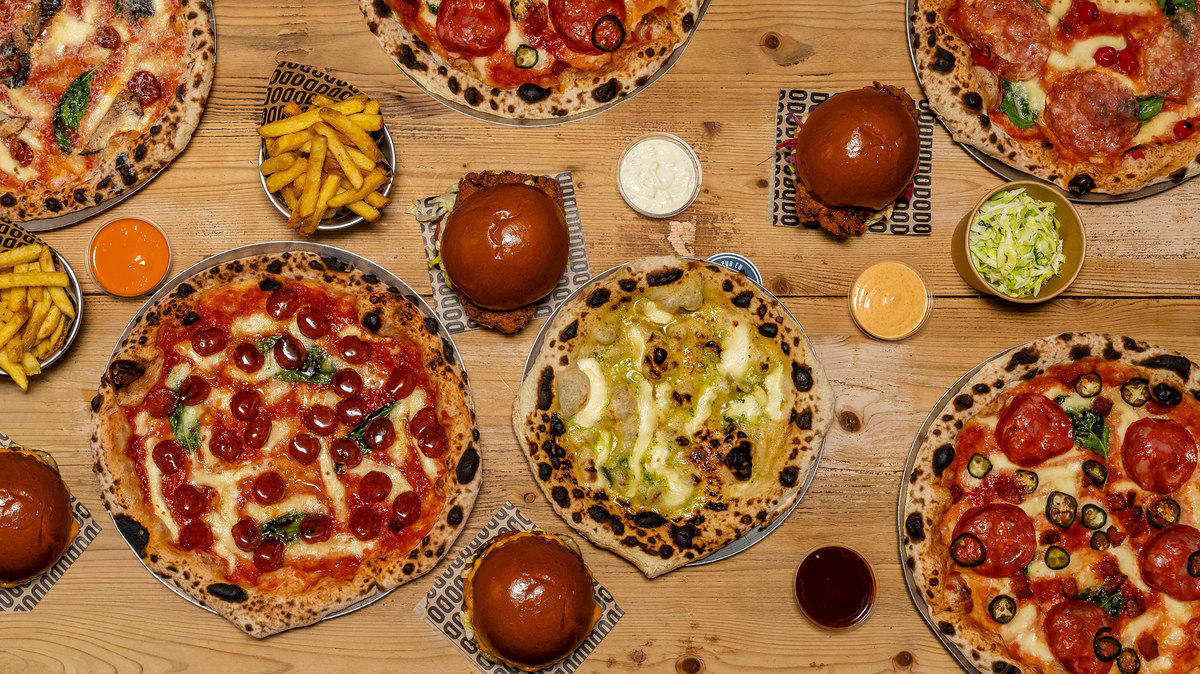 An overhead flat-lay of multiple pizza pizzas on a wooden table, accompanied by fries, small bowls of sauce and toppings, and burger buns.