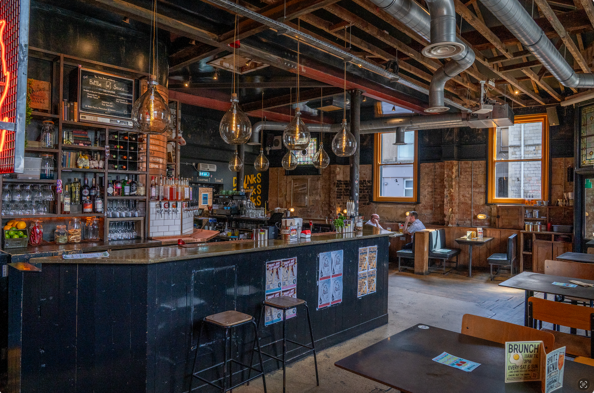 Bar interior with wooden counter, stools, and various bottles on shelves.