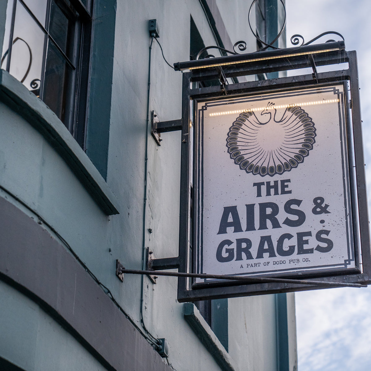 A hanging rectangular sign for 'The Airs & Graces' mounted on the exterior of a building. The sign is white with black lettering and a black decorative emblem at the top, set against a blue sky with clouds.
