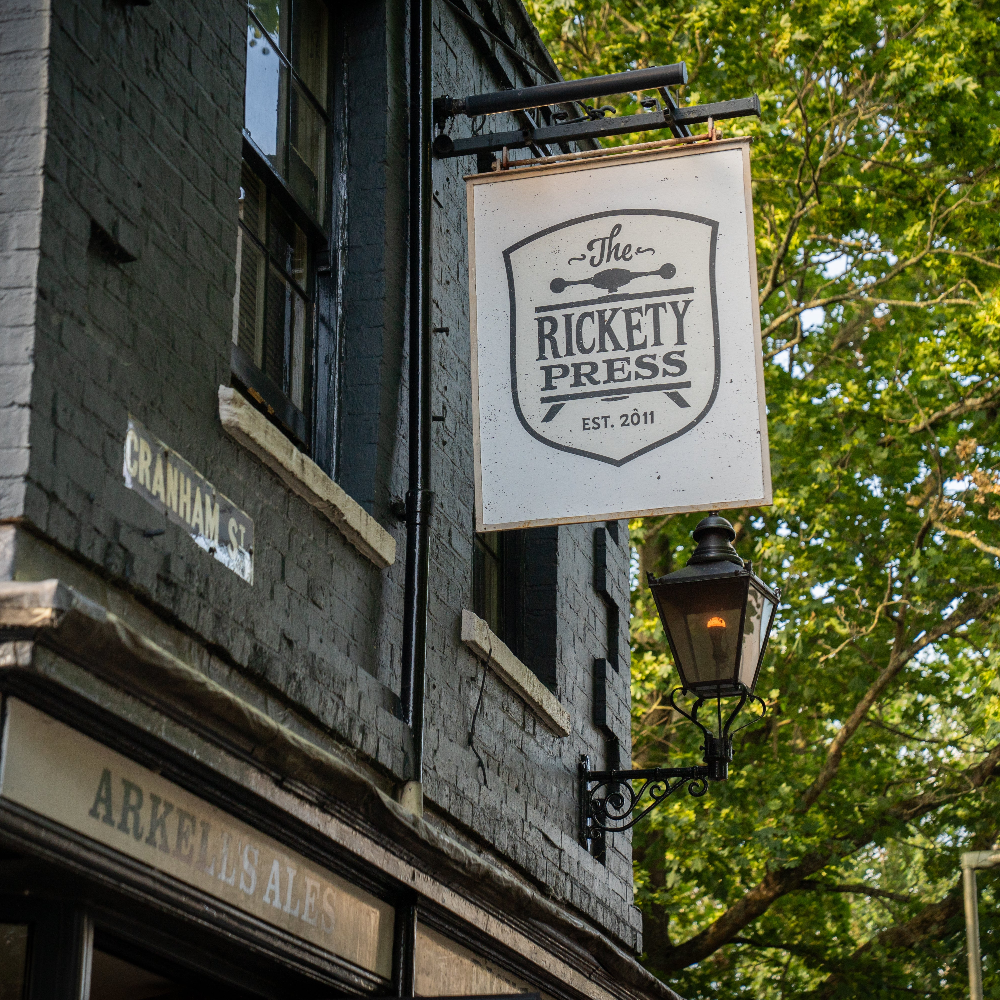 Sign for 'The Rickety Press' on a building exterior with trees in the background