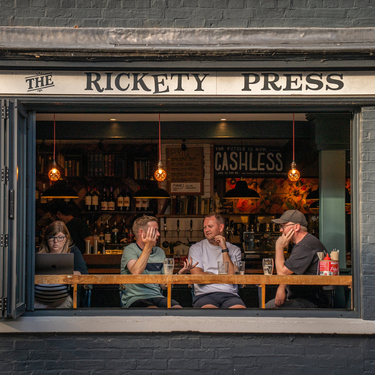 A street-facing view of a dark gray brick building with a large window displaying the interior of 'The Rickety Press' pub. Three men and a woman are seated at a wooden counter inside, and warm hanging bulbs glow from within.