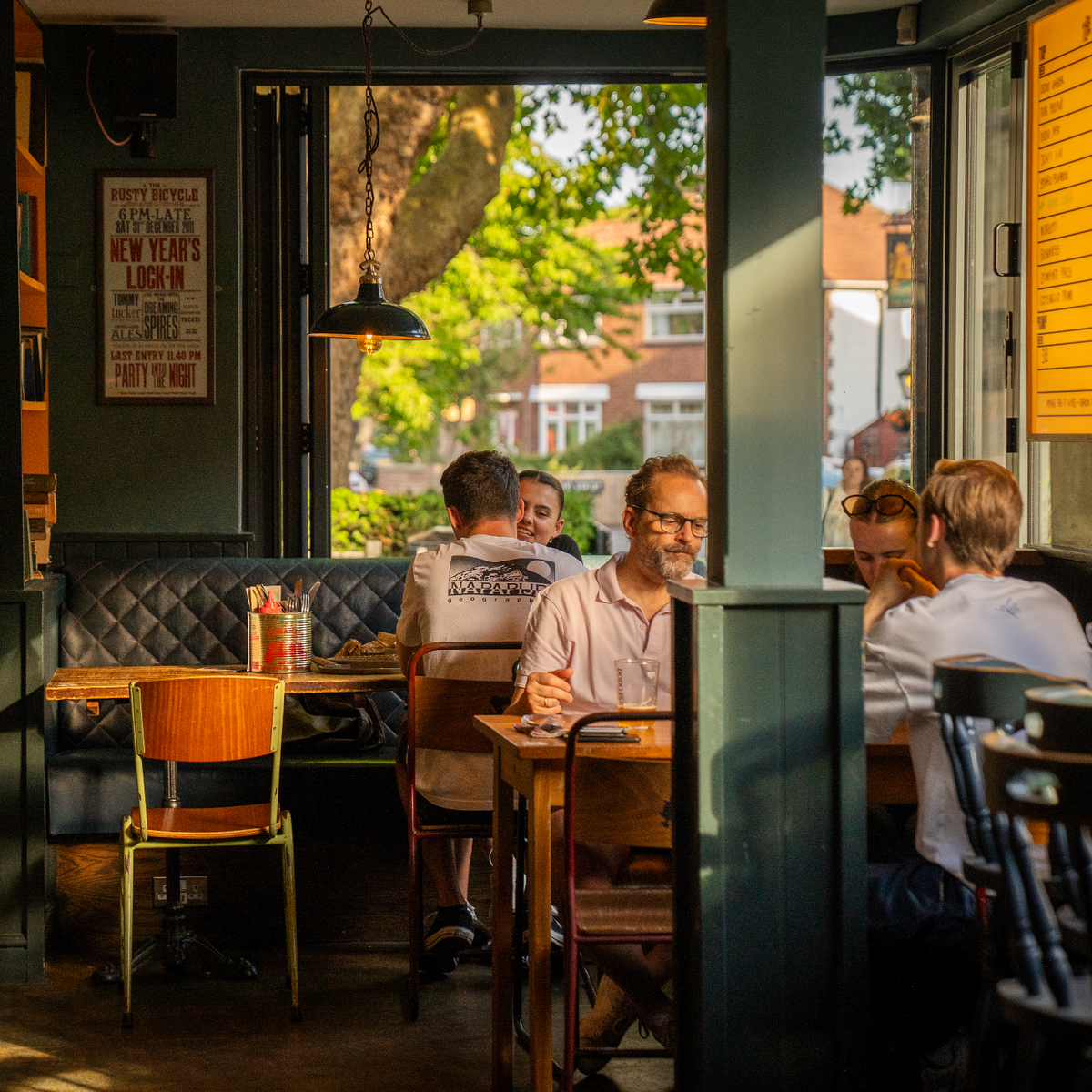 A cozy restaurant interior with several people seated at tables near large windows, with a dark green booth, wooden chairs, and a bright outdoor view of trees and buildings.