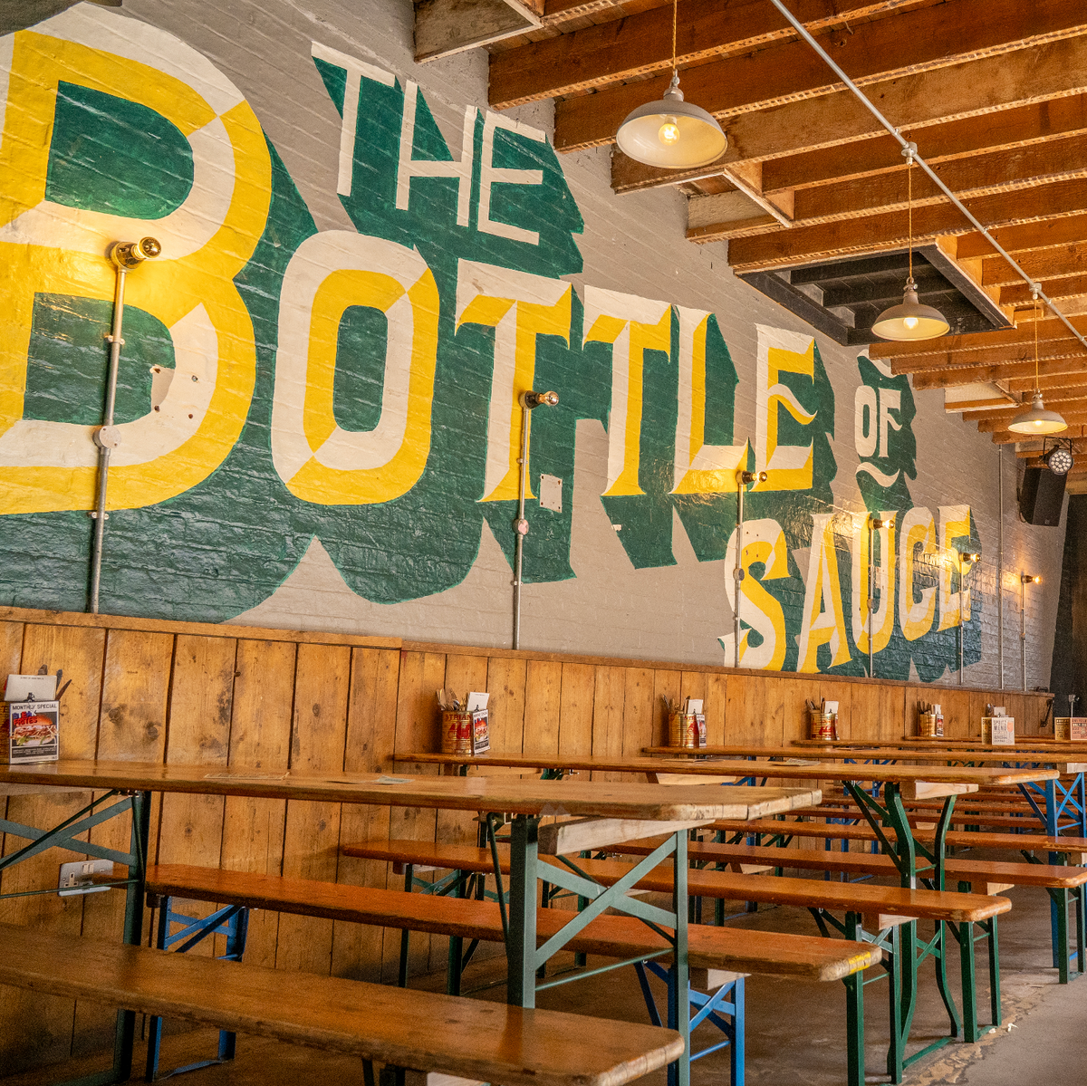 Long wooden tables and benches beneath a large painted The Bottle of Sauce mural