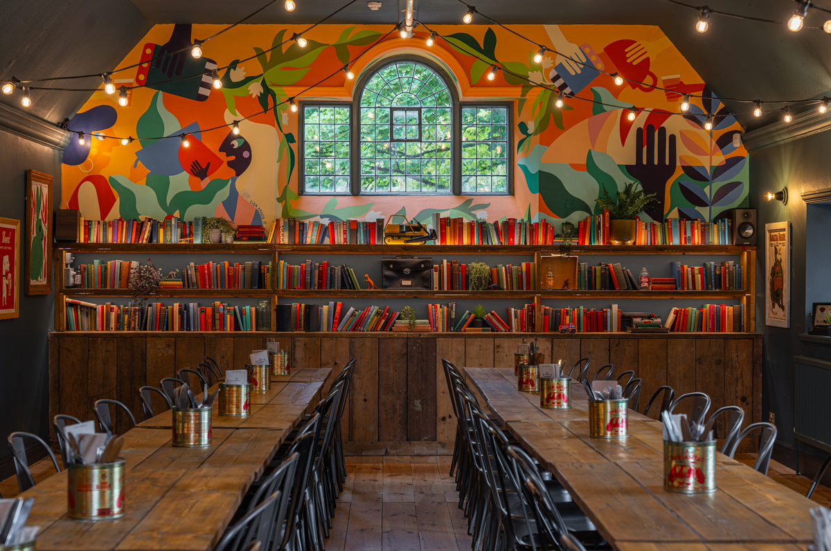 Interior of The Slow & Steady with long tables, bookshelves and a colourful painted mural